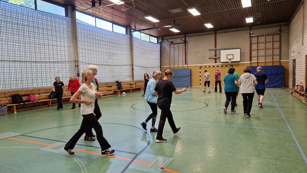 Gruppe Erwachsener bewegt sich gemeinsam durch eine Turnhalle, in lockeren Schritten beim Aufwärmen.“