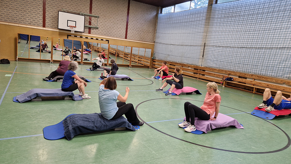 Mehrere Frauen und Männer sitzen auf Matten in einer Sporthalle und machen Bauchmuskelübungen. Im Hintergrund Spiegel, Basketballkorb und Sportgeräte.