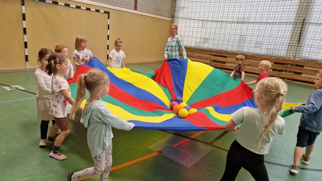 Gruppe von Kindern spielt in der Turnhalle mit einem bunten Schwungtuch, in dessen Mitte bunte Bälle liegen.