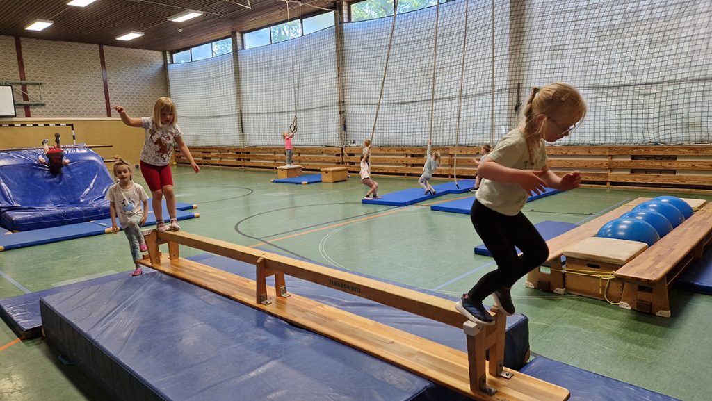 Kinder balancieren, schwingen und turnen an verschiedenen Stationen in einer Turnhalle.