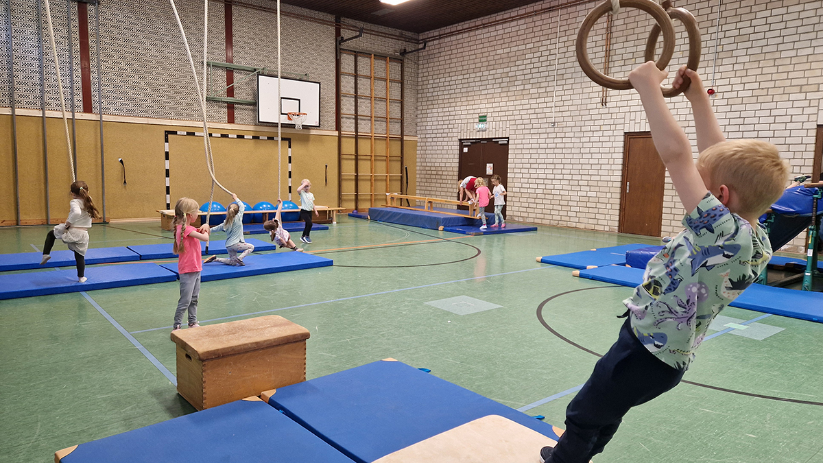 Mehrere Kinder turnen in einer Sporthalle: eines schwingt an Ringen, andere balancieren oder klettern auf Geräten.