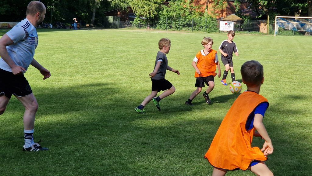 Kinder und ein Erwachsener spielen bei sonnigem Wetter Fußball auf dem Rasenplatz des SV Hertha Otze. Einige Kinder tragen orange Leibchen, andere schwarze Trikots. Der Ball ist in der Luft, alle sind in Bewegung.