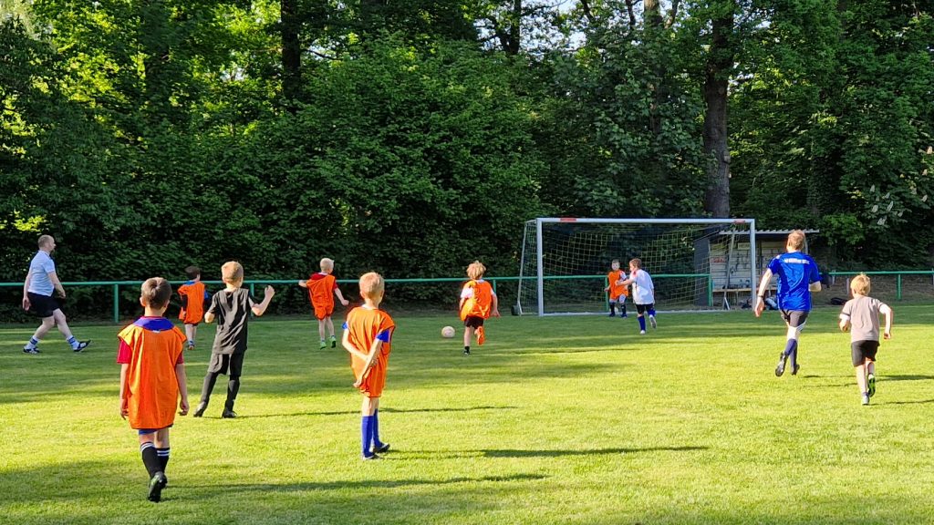 Kinderfußballspiel auf dem Rasenplatz, mehrere Kinder in orangenen Leibchen laufen auf das Tor zu