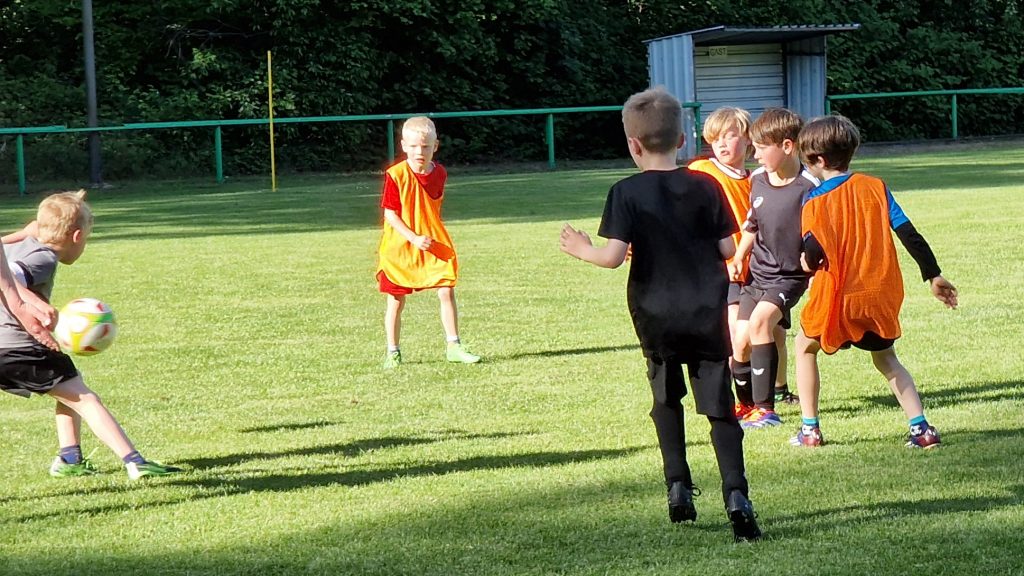 Kinder beim Fußballtraining auf dem Rasenplatz, einige mit orangenen Leibchen, andere in schwarzer Kleidung
