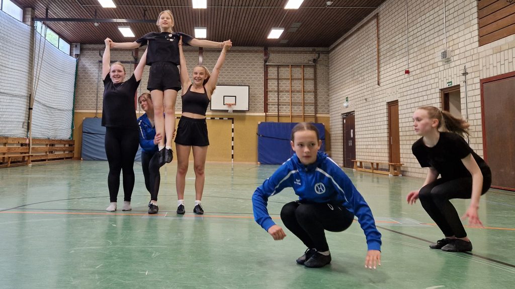 Die Tanzgruppe Diamonds führt in der Turnhalle eine Hebefigur auf, zwei Mädchen heben ein weiteres Mädchen hoch, während vorn zwei Tänzerinnen in Bewegung sind.
