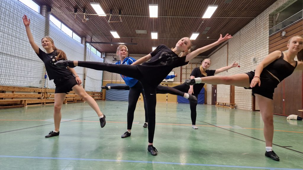 Fünf Tänzerinnen der Gruppe Diamonds stehen in der Turnhalle in einer synchronen Pose mit einem gestreckten Bein zur Seite und erhobenen Armen.