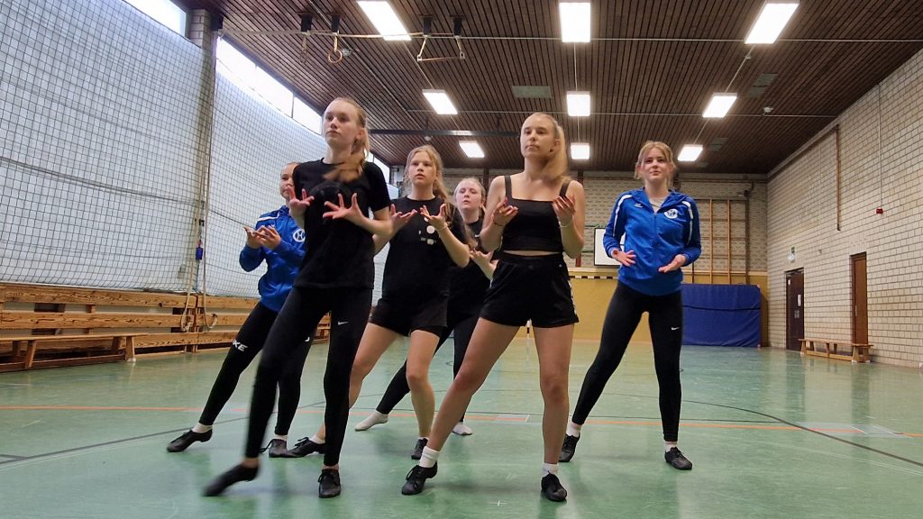 Sechs Tänzerinnen der Gruppe Diamonds stehen in Formation mit leicht gebeugten Armen und offenen Handflächen in Richtung Gesicht – eine ausdrucksstarke Tanzpose in der Sporthalle.