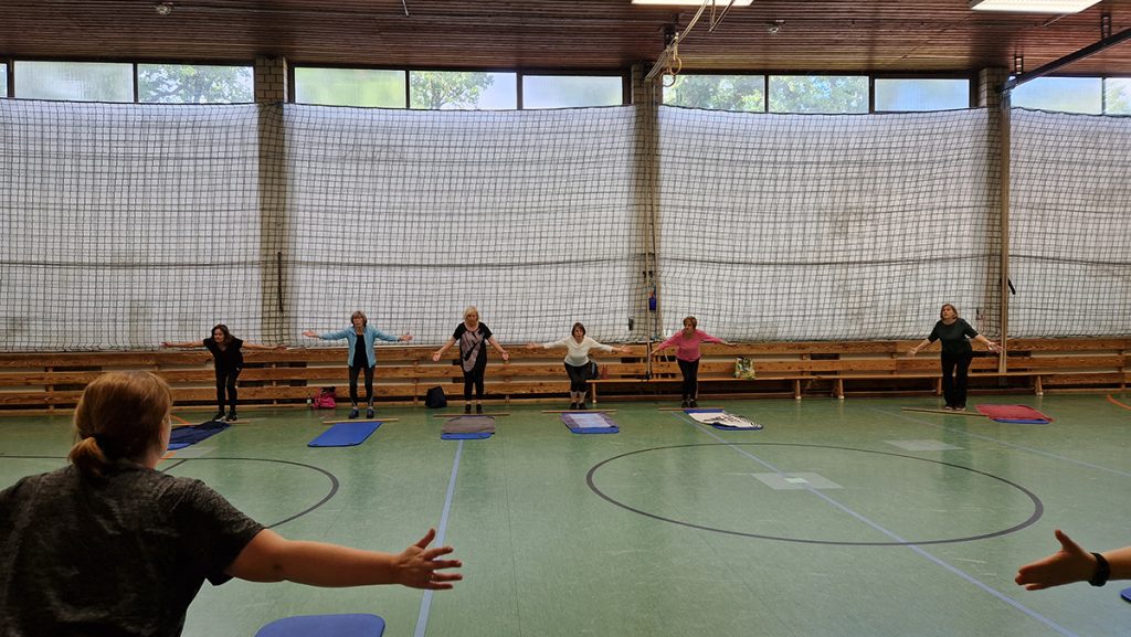 Mehrere Erwachsene stehen auf Gymnastikmatten in einer Turnhalle mit ausgestreckten Armen. Die Gruppe macht gemeinsam Rückengymnastik, im Hintergrund sind Bänke und ein Netz an der Wand zu sehen.