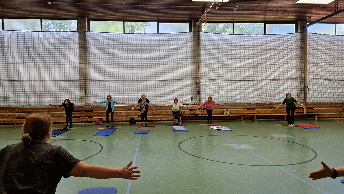 Mehrere Erwachsene stehen auf Gymnastikmatten in einer Turnhalle mit ausgestreckten Armen. Die Gruppe macht gemeinsam Rückengymnastik, im Hintergrund sind Bänke und ein Netz an der Wand zu sehen.