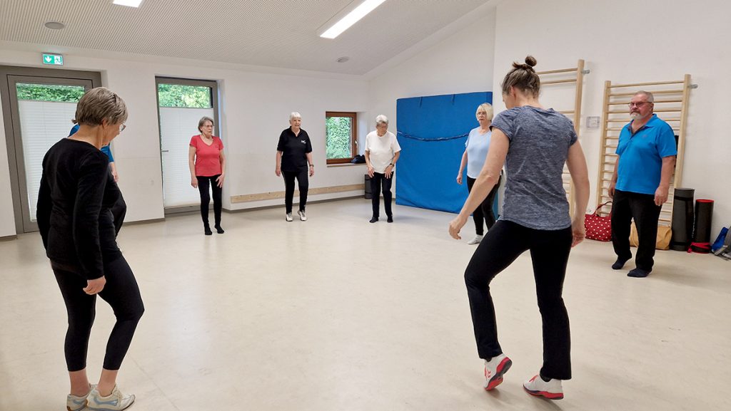 Gruppe älterer Menschen steht im Kreis im Bewegungsraum der KiTa Otze. Eine Trainerin zeigt eine Bewegung mit den Beinen, die Teilnehmenden folgen der Übung.