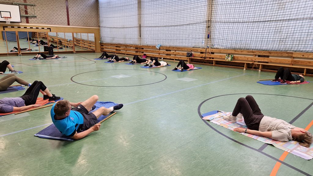 Mehrere Erwachsene liegen in einer Turnhalle auf Gymnastikmatten und führen gemeinsam eine Rückenübung in Rückenlage aus.