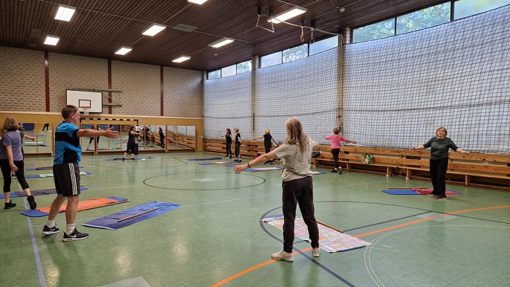 Erwachsene führen gemeinsam in einer Turnhalle eine stehende Übung zur Mobilisierung und Kräftigung durch.