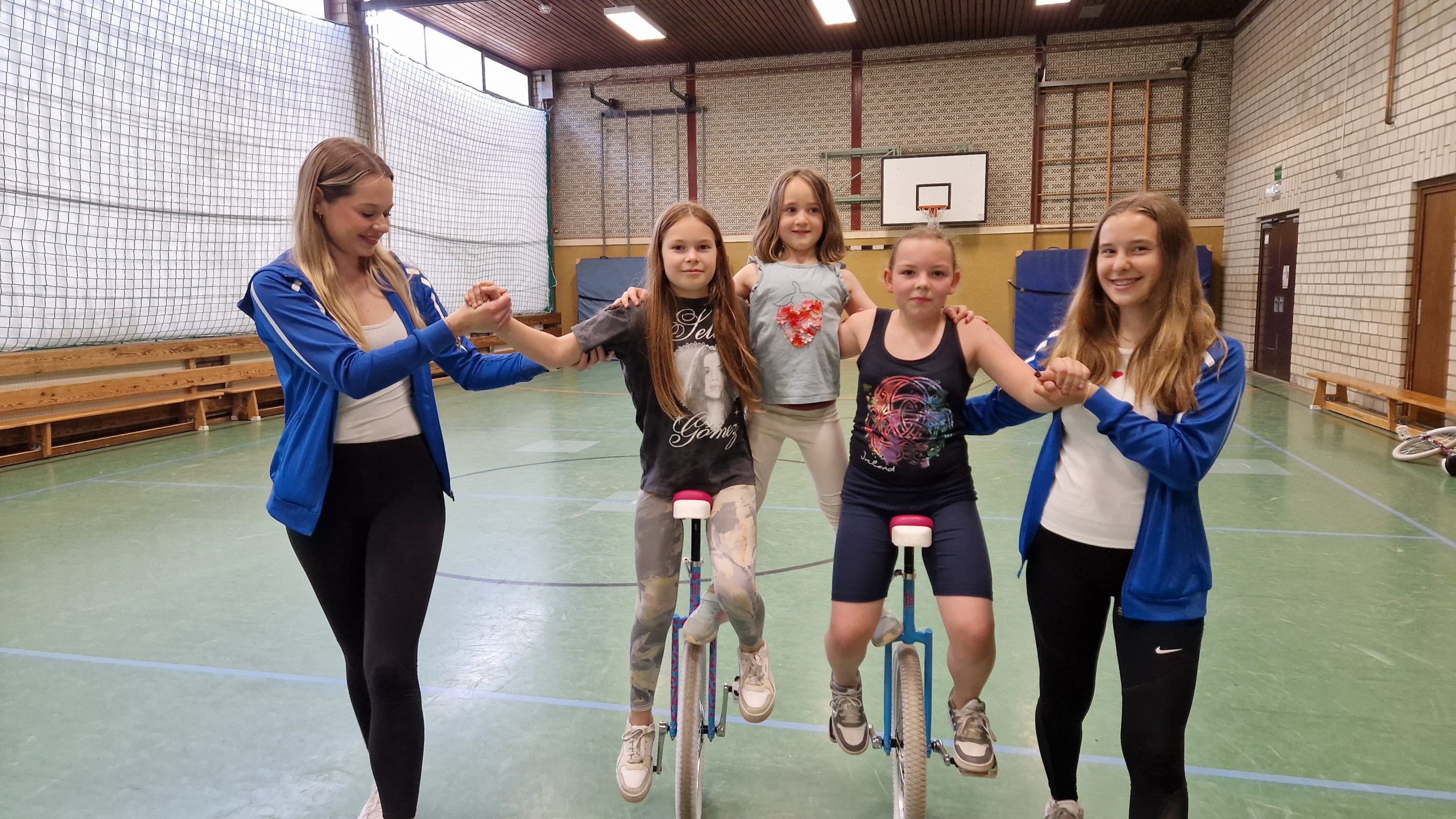 Zwei Mädchen fahren in einer Sporthalle Einrad und werden links und rechts von zwei älteren Mädchen gesichert. Ein weiteres Kind steht zwischen ihnen. Alle lächeln und halten sich an den Händen.