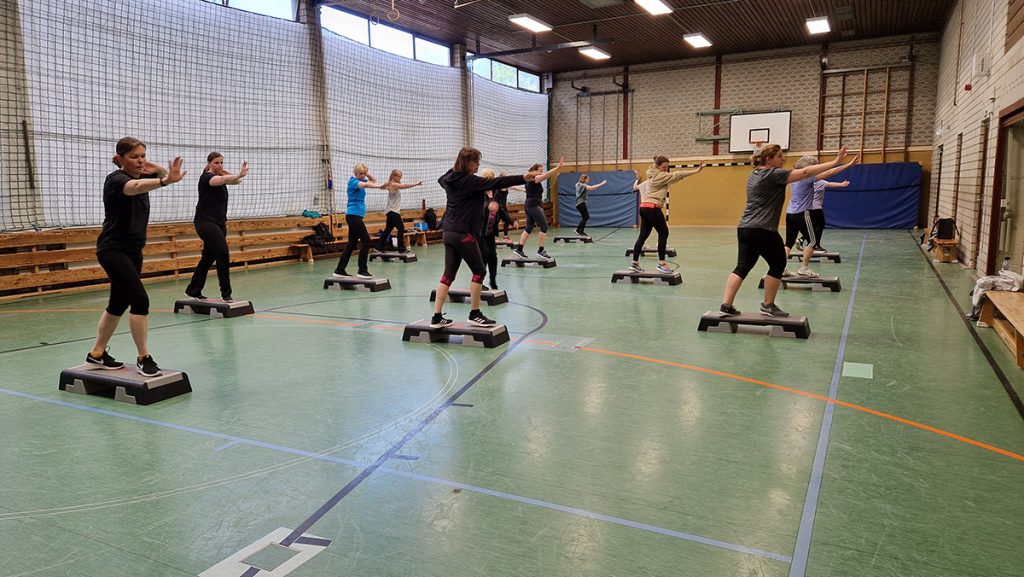 Teilnehmerinnen einer Step-Aerobic-Gruppe beim Training in einer Sporthalle. Jede Person steht auf einem Step-Brett und führt synchron eine Armbewegung nach vorne aus.