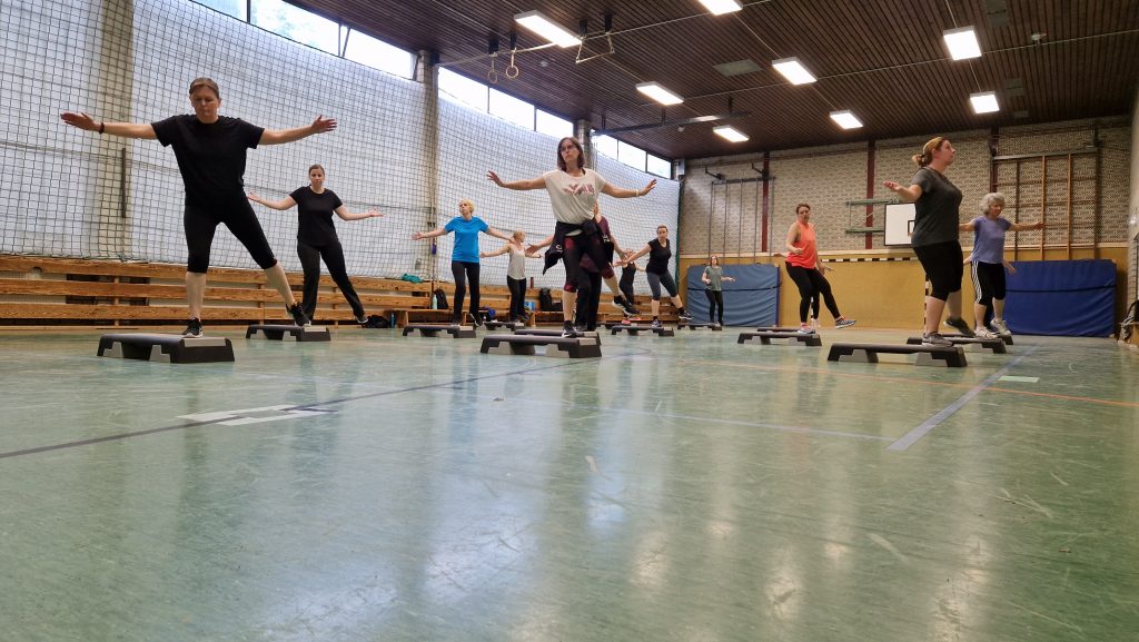 Step-Aerobic-Gruppe in einer Turnhalle beim Training. Die Teilnehmenden stehen mit ausgestreckten Armen auf Step-Brettern und führen eine koordinierte Übung aus.