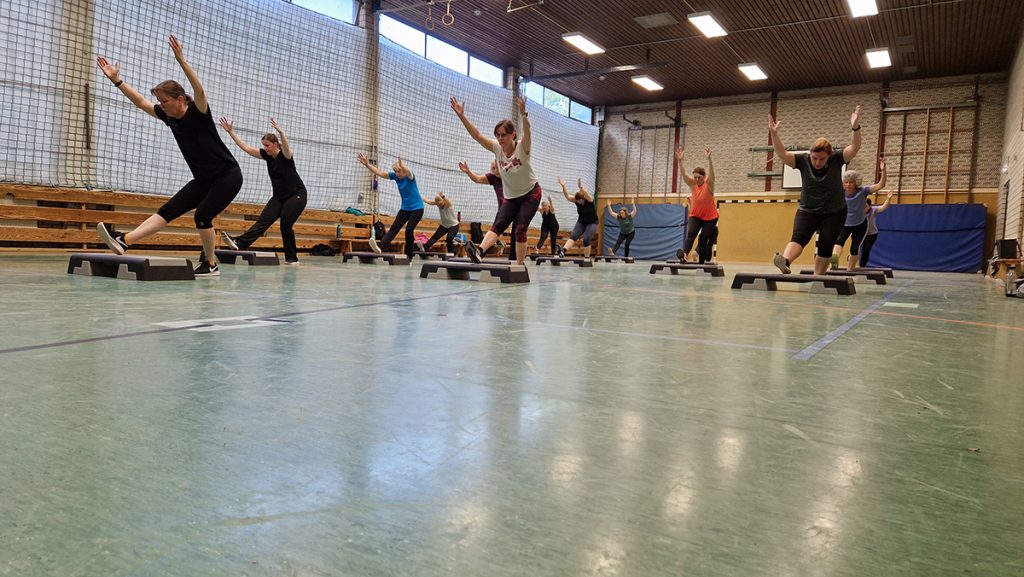 Teilnehmerinnen einer Step-Aerobic-Gruppe in einer Turnhalle heben gleichzeitig die Arme und ein Bein, während sie auf Step-Brettern stehen. Die Übung betont Koordination und Gleichgewicht.