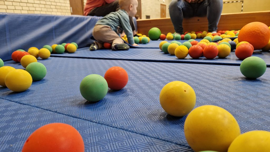 Ein Kleinkind krabbelt auf einer blauen Matte, umgeben von vielen bunten Softbällen. Im Hintergrund sitzen zwei Erwachsene und beobachten die Szene aufmerksam.