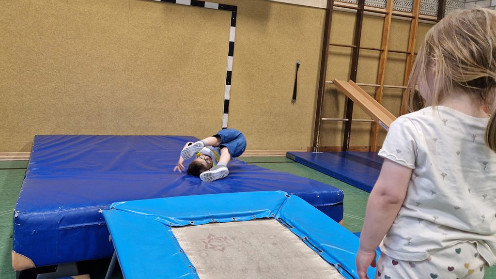 Ein Kind landet nach einem Sprung vom Trampolin auf einer großen blauen Matte, ein weiteres Kind beobachtet die Szene.