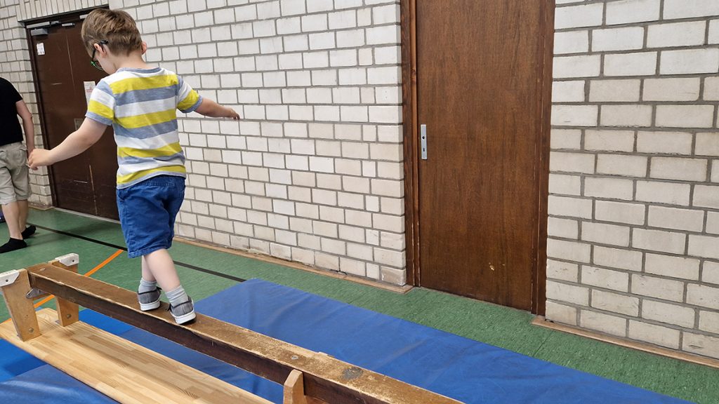 Ein Kind balanciert konzentriert über eine schmale Holzbank in der Turnhalle, darunter liegt eine Matte.