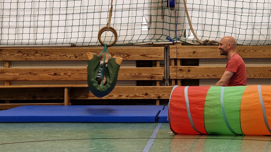 Ein Kleinkind schaukelt sicher in einer grünen Mattenchaukel an einem Turnring, während ein lachender Erwachsener daneben sitzt. Im Vordergrund liegt ein bunter Krabbeltunnel.