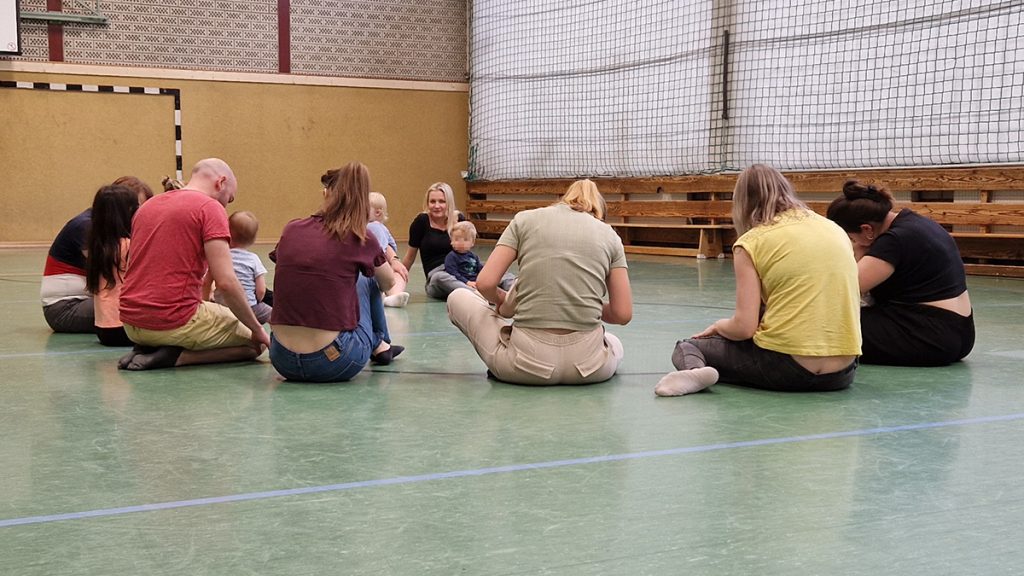 Erwachsene sitzen mit ihren Kleinkindern im Kreis auf dem Hallenboden. Die Atmosphäre ist ruhig und konzentriert.
