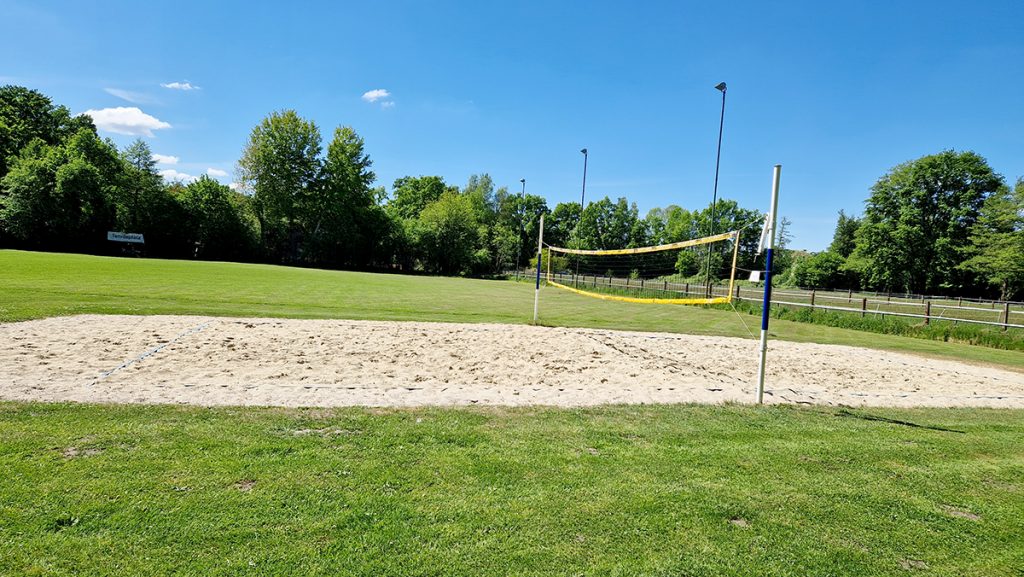 Beachvolleyballfeld auf einer sonnigen Wiese mit Netz und Sandfläche, im Hintergrund Bäume und ein grüner Sportplatz.