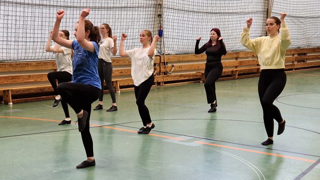 Sechs junge Frauen der Blond AG tanzen synchron mit erhobenen Armen und angehobenem Bein in der Sporthalle.