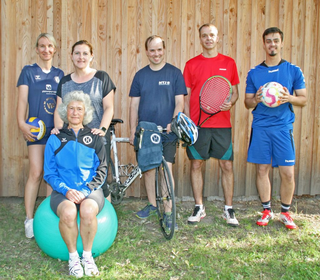Sechs Erwachsene des SV Hertha Otze stehen vor einer Holzfassade und präsentieren ihre Sportarten: Volleyball, Tanzen, Gymnastik, Triathlon, Tennis und Fußball.