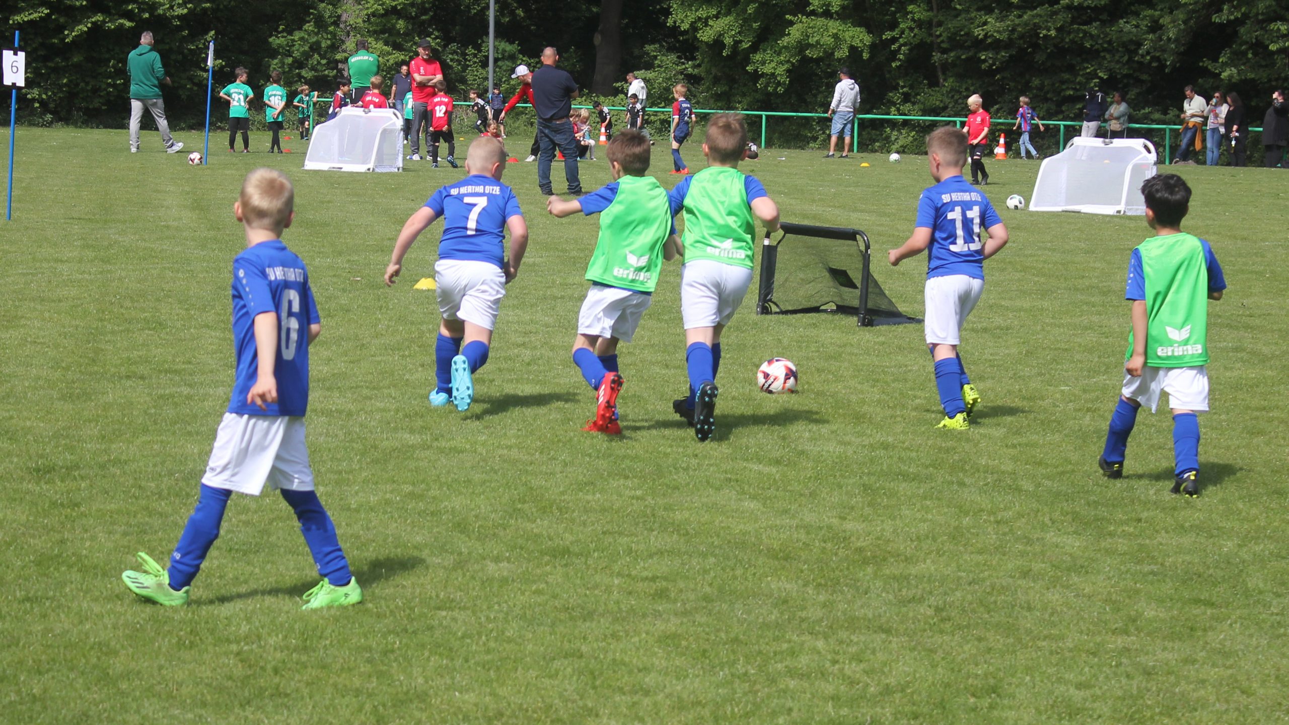 Mehrere Kinder der G-Jugend des SV Hertha Otze spielen auf dem Fußballfeld. Einige tragen blaue Trikots, andere grüne Leibchen. Im Hintergrund sind kleine Tore und Zuschauer zu sehen.