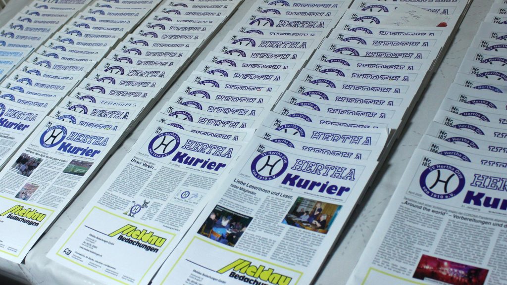 Stapelweise ausgelegte Vereinszeitungen „Hertha Kurier“ mit blauem Logo des SV Hertha Otze und gelben Werbeanzeigen.