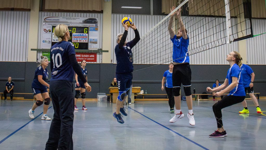 Spielszene eines Volleyballspiels in der Halle zwischen einem Frauen- und einem Männerteam – eine Spielerin springt zum Block gegen zwei Gegenspieler.
