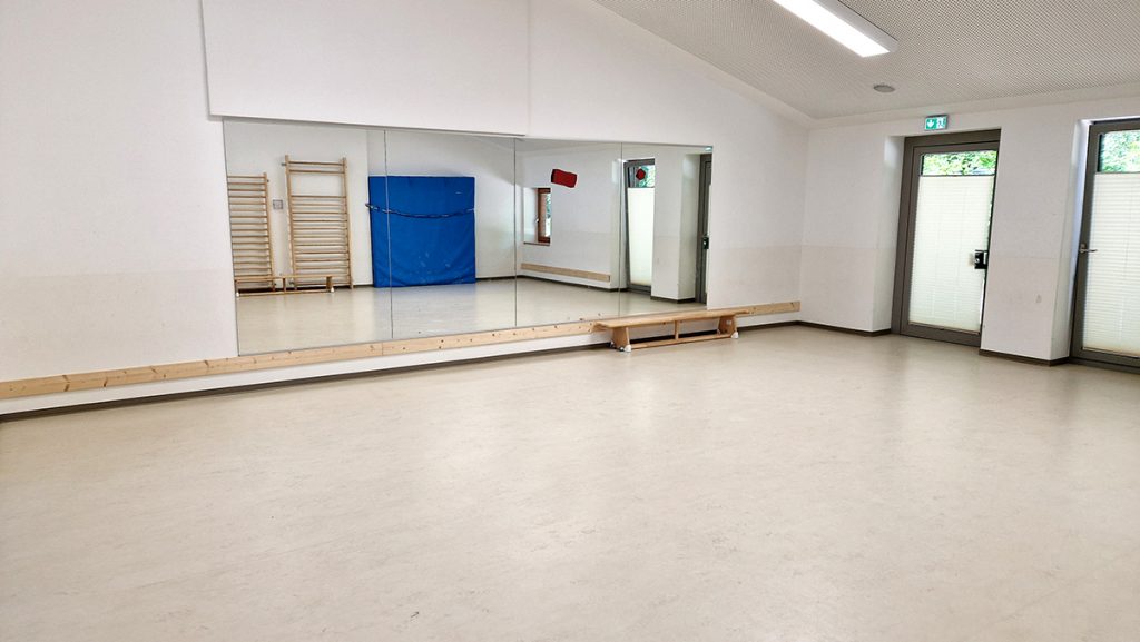 Heller Bewegungsraum mit großem Spiegel an der Wand, Sprossenwand, Turnmatte und bodentiefen Fenstern mit Sichtschutz. Der Raum ist leer und bereit für sportliche Aktivitäten.