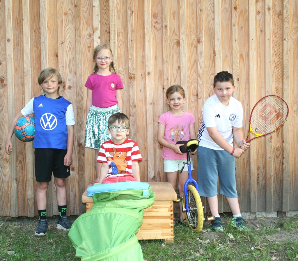 „Fünf Kinder des SV Hertha Otze stehen vor einer Holzfassade und zeigen ihre Sportarten: Fußball, Tanzen, Kinderturnen, Einrad und Tennis.