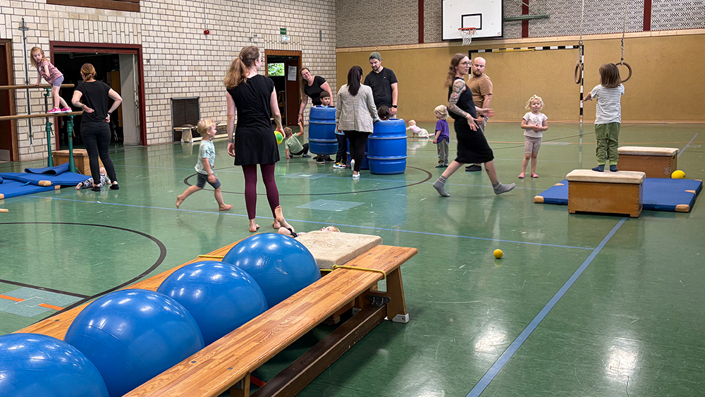 Kinderturnen in der Sporthalle – Kleinkinder mit Eltern an verschiedenen Stationen.