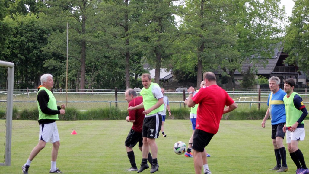Erwachsene Männer verschiedenen Alters spielen Fußball auf einem Rasenplatz. Einige tragen farbige Leibchen, andere Trikots. Im Vordergrund wird der Ball gerade gespielt, während alle aktiv am Spiel teilnehmen.