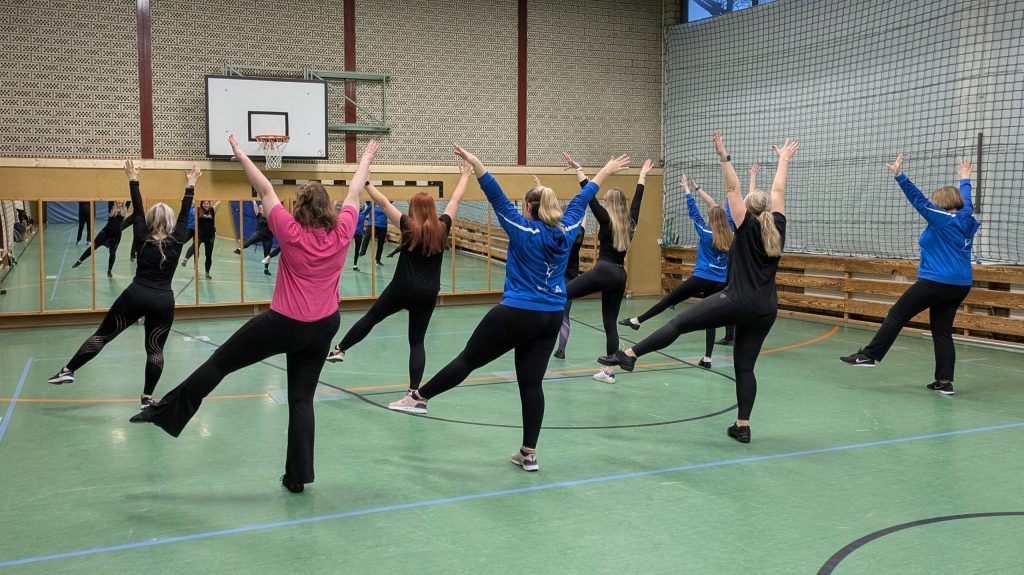 Gruppe von Frauen beim Tanzfitness-Training in einer Sporthalle. Alle heben synchron Arme und Beine – im Vordergrund eine Frau in pinkem Shirt, mehrere tragen blaue Vereinsjacken.