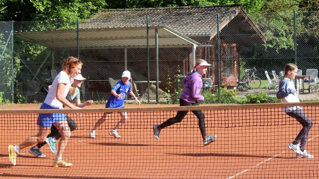 Sechs Spielerinnen, darunter eine Trainerin, machen Laufübungen auf dem Tennisplatz. Alle bewegen sich gleichzeitig in Laufrichtung auf dem roten Sandplatz.