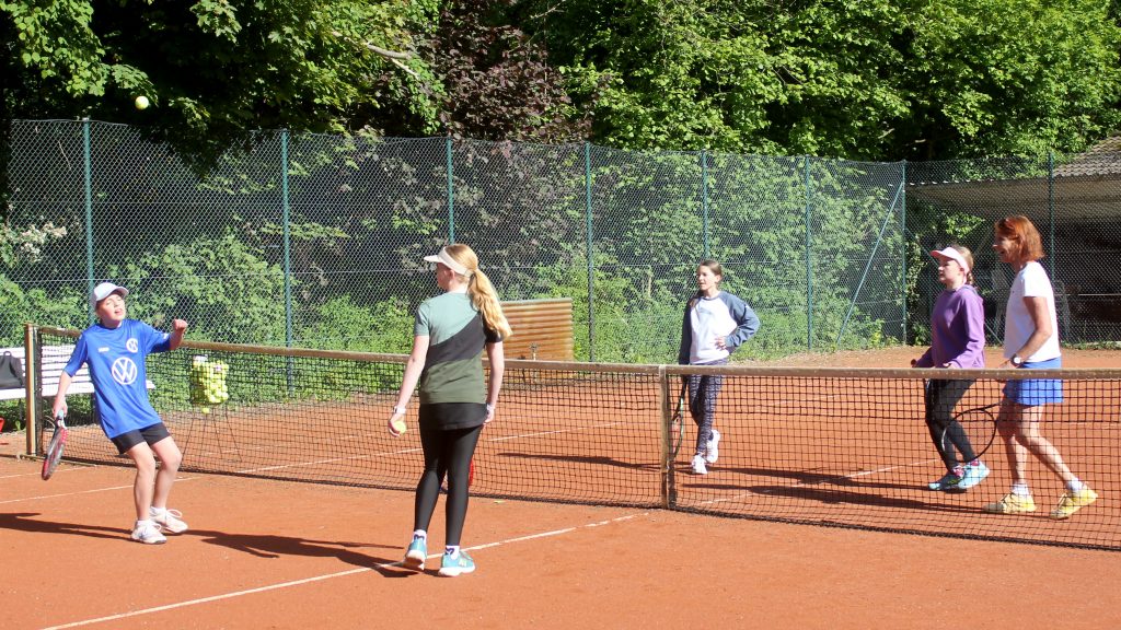 Fünf Personen stehen auf einem Tennisplatz verteilt: Zwei auf der linken und drei auf der rechten Seite des Netzes. Alle beobachten einen Tennisball, der hoch über dem Netz fliegt.
