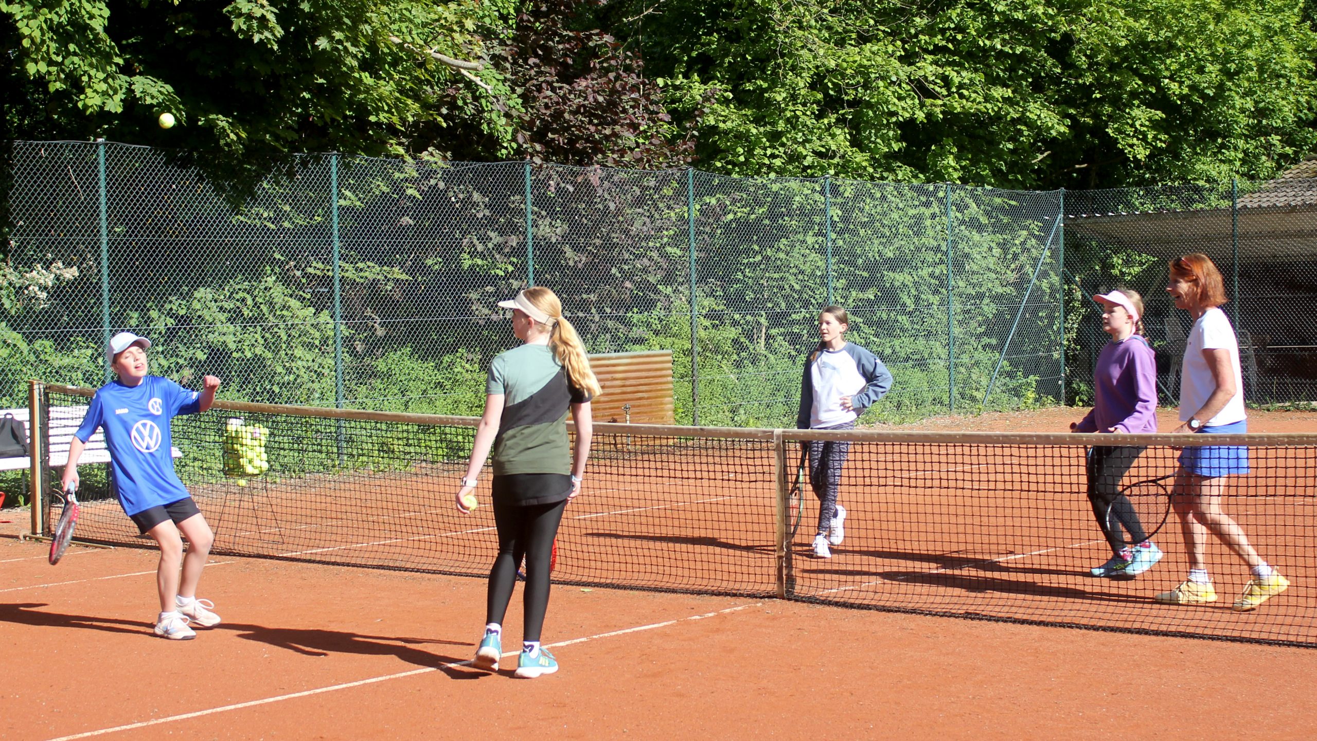 Fünf Personen stehen auf einem Tennisplatz verteilt: Zwei auf der linken und drei auf der rechten Seite des Netzes. Alle beobachten einen Tennisball, der hoch über dem Netz fliegt.