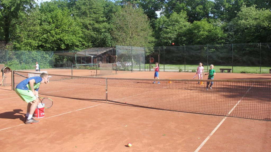 Kinder trainieren mit Tennisschlägern auf einem Sandplatz unter Anleitung eines Trainers.