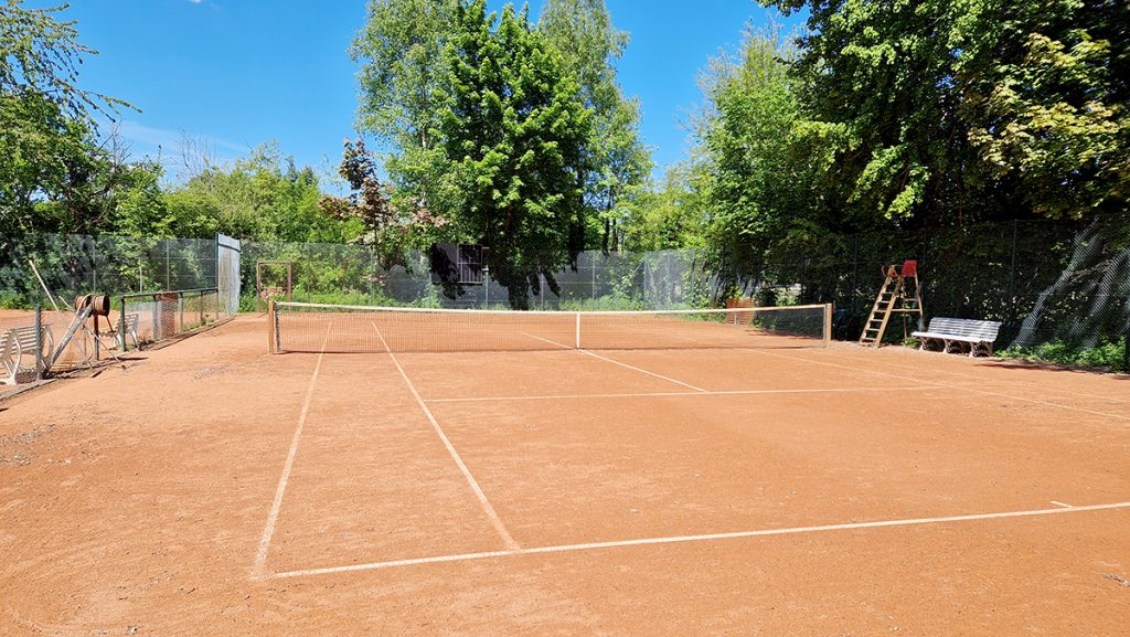 Tennisplatz mit rotem Sandbelag und Netz, umgeben von hohen Bäumen und Zäunen.