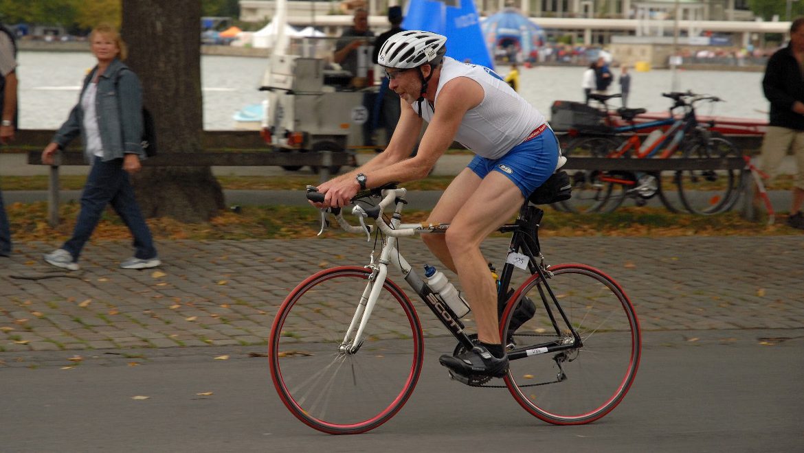 Triathlet mit Helm und Rennrad fährt auf einer Straße am Wasser entlang, im Hintergrund sind Zuschauer, Fahrräder und Veranstaltungszelte zu sehen.
