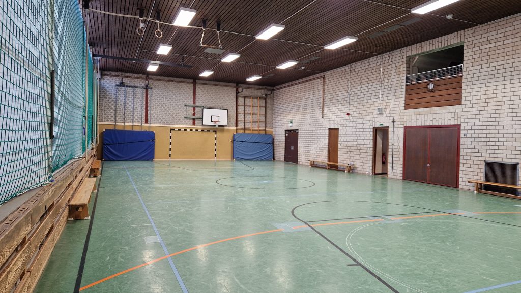 Leere Turnhalle mit grünem Boden, Basketballkorb, Kletterwand und gepolsterten Wänden.