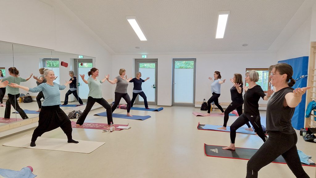 Frauen stehen barfuß auf Yogamatten im Bewegungsraum der KiTa Otze. In einer Halbkreisformation machen sie unter Anleitung eine stehende Yoga-Haltung mit ausgestreckten Armen. Im Hintergrund Spiegel und große Fenster.